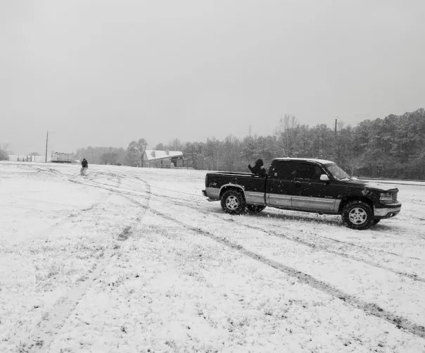 Someone is pulled by a snowboard behind a truck. The truck is in the foreground.