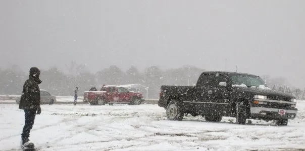 Someone on waits on a snowboard in the foregronud a black and red truck in the distance.