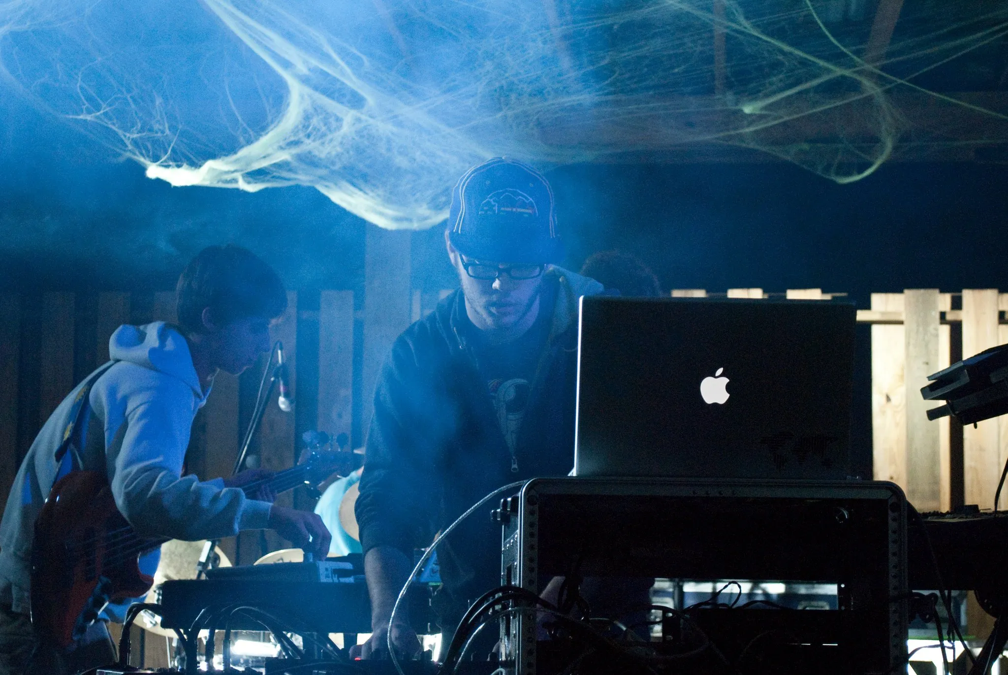 A DJ plays behind is macbook against a smokey blue background.