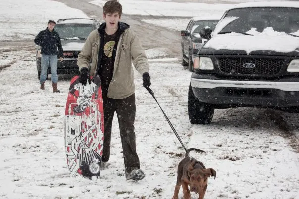 Luke holds a boogie board while his dog pulls his leash in the snow.