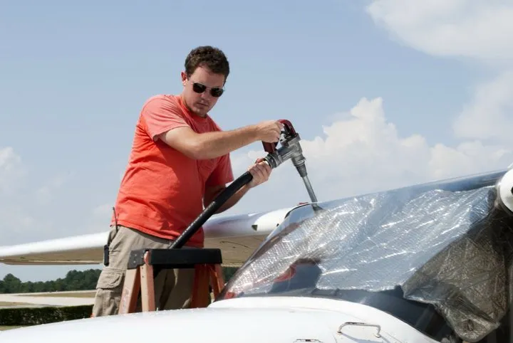 A guy in a red shirt with aviators stands on a ladder to fuel a small prop plane.