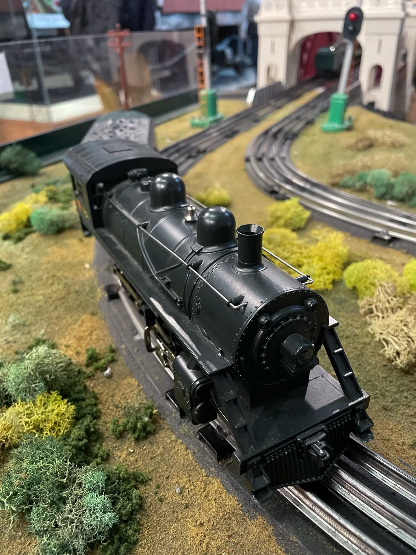 A detailed model train, a black Lionel locomotive, sits on a miniature track surrounded by realistic scenery. The train model, with intricate designs and a glossy finish, captures the essence of a bygone era of steam engines. The landscape includes textured greenery resembling moss and small shrubs, adding depth and life to the miniature world. In the background, model railway signals and a tunnel entrance enhance the illusion of a bustling model train set environment.