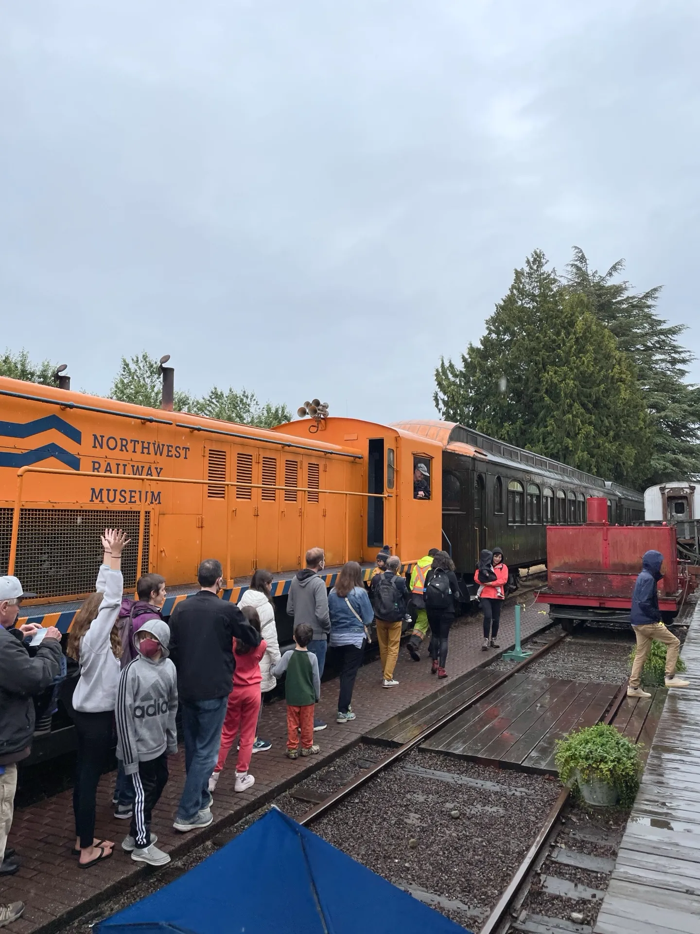 A group of visitors, including children and adults, gather around a bright orange locomotive at the Northwest Railway Museum. They appear engaged and excited, with some waving towards the train engineer. The vibrant colors of the train stand out against the overcast sky.