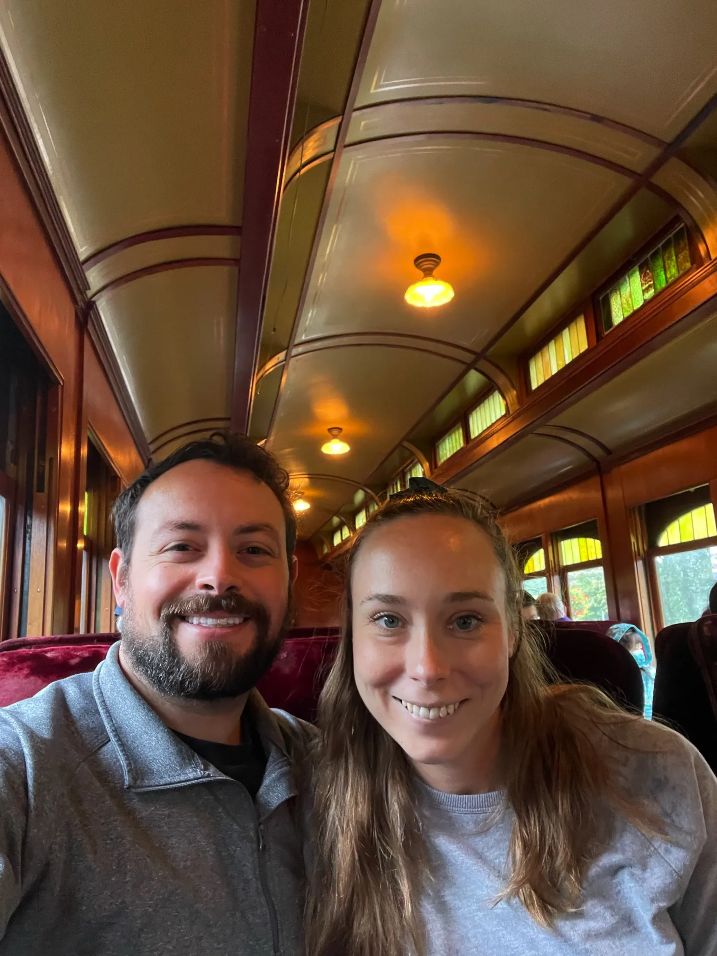 A selfie of us on a vintage train ride. The rich wooden interior of the train carriage, complete with maroon velvet seats and warm lighting, adds a classic touch to the photo. Stained glass windows on the upper part of the carriage allow a hint of natural light to seep in, enhancing the nostalgic ambiance of their adventure."