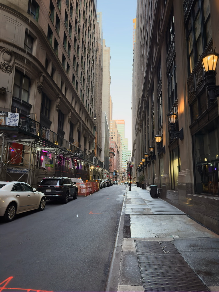 A narrow city street lined with tall buildings and warm wall-mounted lights.